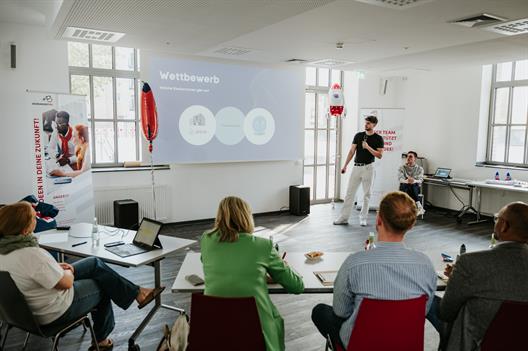 Das Bild zeigt, wie ein junger Mann seine Gründungsidee mit einer Präsentation vor einer Jury pitcht.